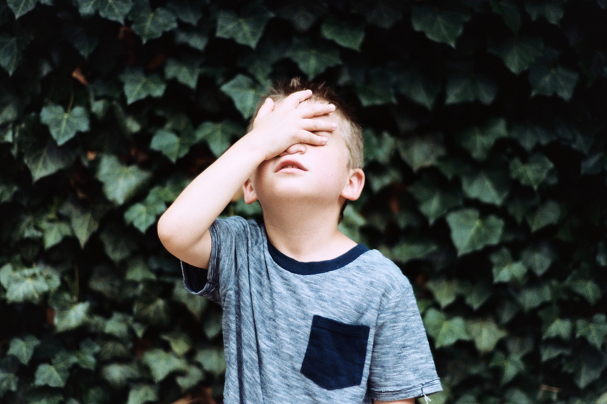 image of a young boy with his hand over his face in annoyance in front of a bunch of leaves