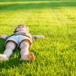 young boy with bare feet lays on back on green lawn