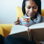 woman on couch reads book and drinks tea from mug