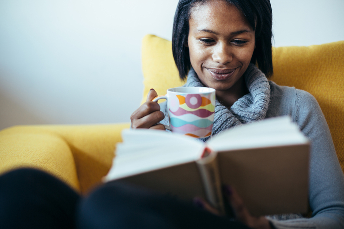 woman on couch reads book and drinks tea from mug