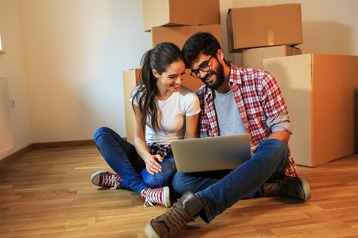 young smiling couple sits with moving boxes and look at laptop