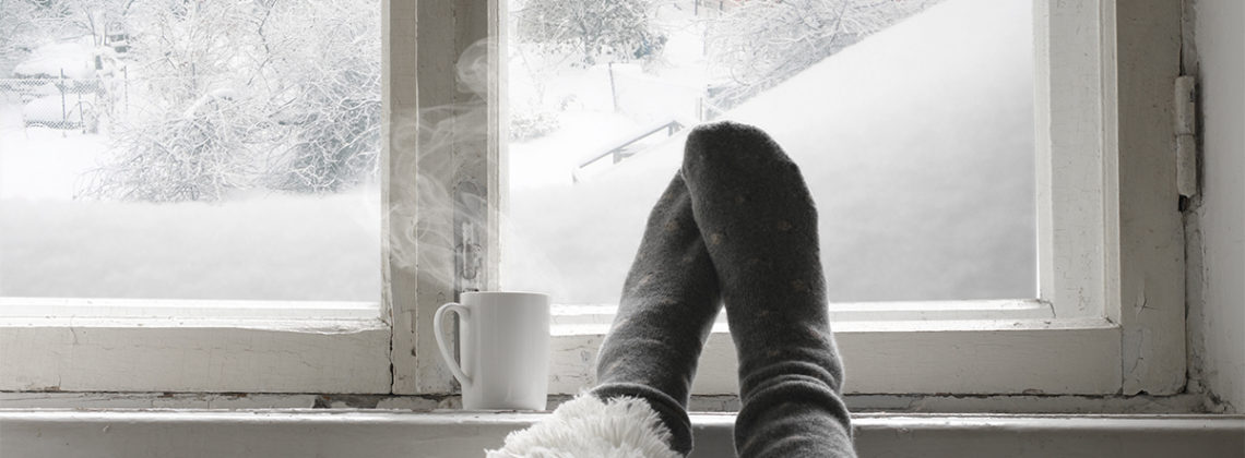 a persons feet with socks in front of a cold window and a steaming mug of liquid