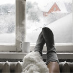 a persons feet with socks in front of a cold window and a steaming mug of liquid