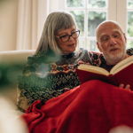 elderly couple sitting on the couch reading a book with a blanket