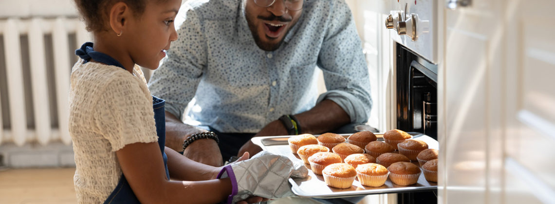little girl pulls muffins from oven while happy father watches