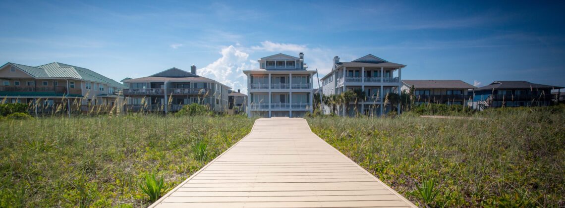 A boardwalk leads through sand dunes to large beach houses.