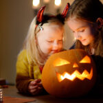two young girls smile at a lit jack-o-lantern on halloween