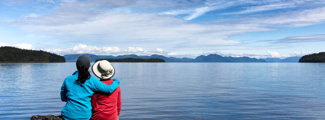 Daughter embraces her mother while they sit before an alpine lake with snowy mountains in the backgroud