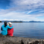 Daughter embraces her mother while they sit before an alpine lake with snowy mountains in the backgroud