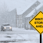 Car driving down a snowy street in a winter storm with houses in the background